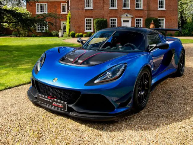 Een blauwe Lotus Exige Cup 380 geparkeerd voor een groot huis.