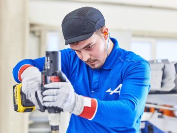 Een man aan het werk met een boormachine in een fabriek, waar hij een Alpine A110 bouwt.