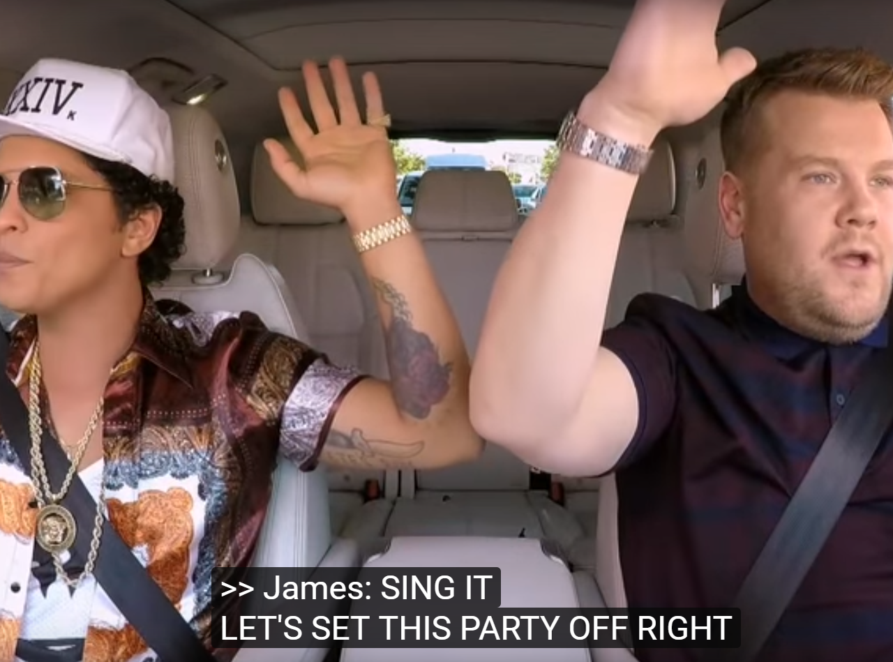Twee mannen in een auto zwaaien met hun handen in de lucht tijdens Carpool Karaoke.