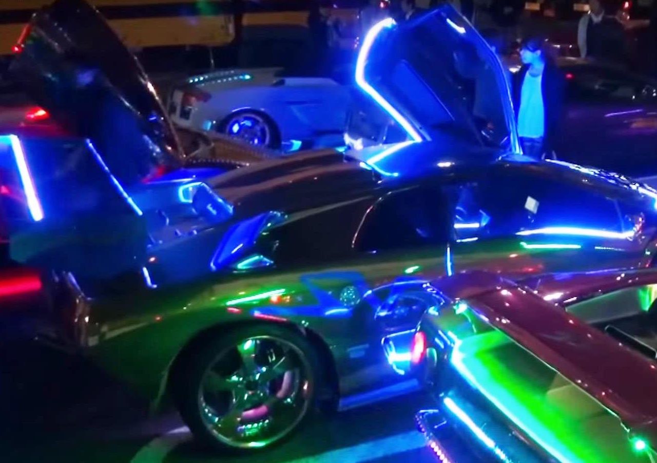 Een groep auto's, waaronder Lamborghini-rijders, parkeerde 's nachts op een neonverlichte parkeerplaats.