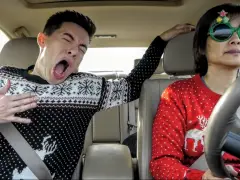 Een man en een vrouw in een auto, gekleed in kersttruien, nemen een hilarische playbackshowvideo op.