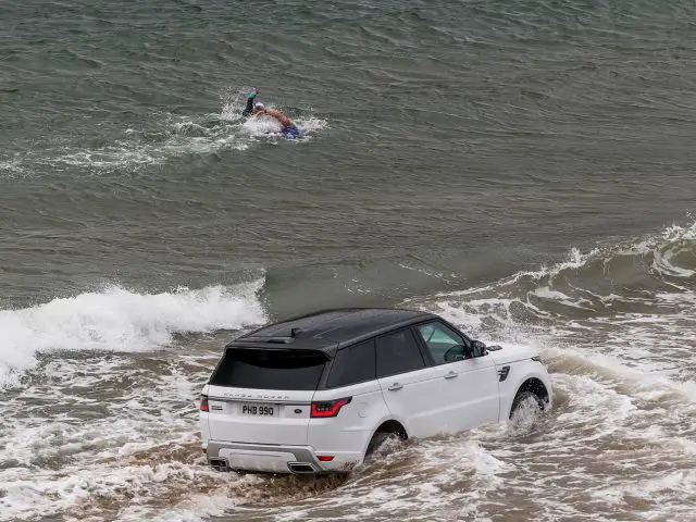 Een witte Range Rover Sport P400e in de oceaan met een surfer in het water.