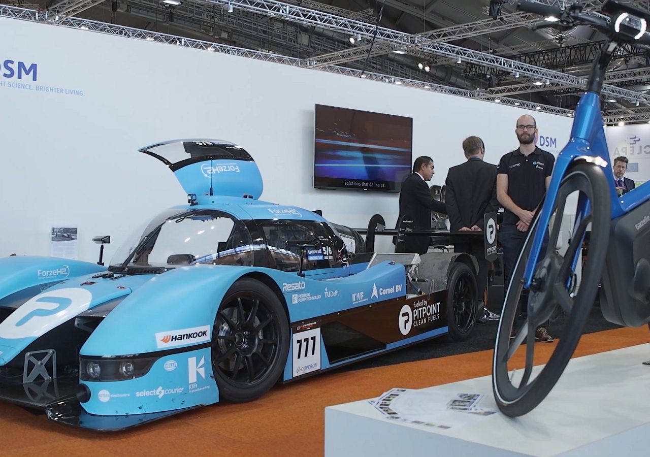 Op een beurs zijn een blauwe raceauto en een fiets te zien, uitgelicht in de videocolumn van de RAI Vereniging.