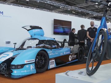 Op een beurs zijn een blauwe raceauto en een fiets te zien, uitgelicht in de videocolumn van de RAI Vereniging.