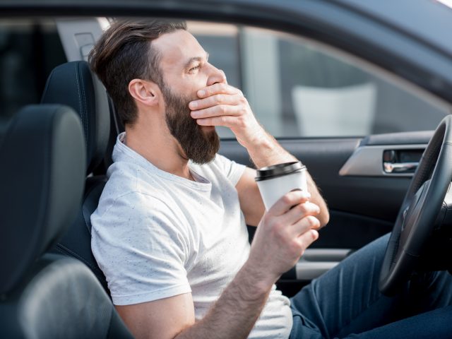 Een slaperige man zit in een auto met een kopje koffie in zijn mond.