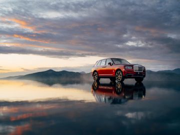 Een rode Rolls-Royce Cullinan geparkeerd op een meer bij zonsondergang.