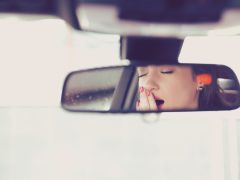 Een vrouw kijkt slaperig naar zichzelf in de achteruitkijkspiegel van een auto.