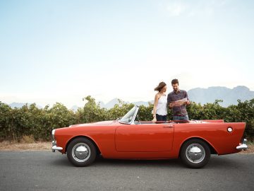 Een man en een vrouw zitten in een rode cabriolet, terwijl de man navigeert.