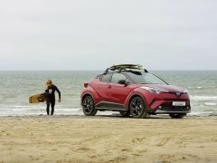 Een rode Toyota C-HR Adventure met een surfplank op het strand.