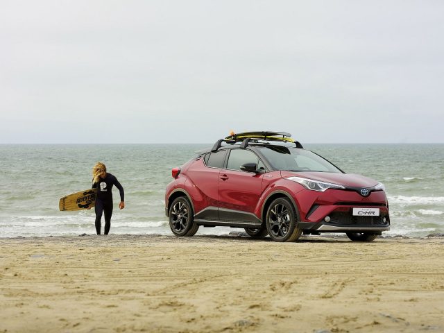 Een rode Toyota C-HR Adventure met een surfplank op het strand.