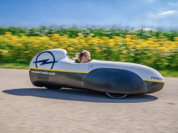 Een vrouw in een Opel die over een weg rijdt met zonnebloemen op de achtergrond.