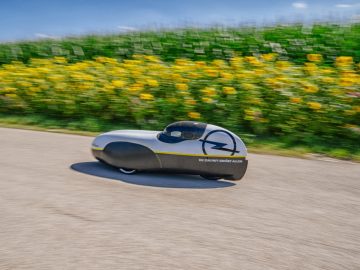 Een kleine Opel-auto rijdt over een weg met zonnebloemen op de achtergrond.