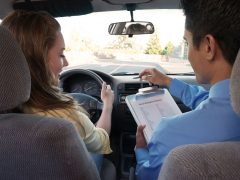 Een man en een vrouw in een auto met een klembord in de hand, pratend over de prijzen voor hun rijbewijs.