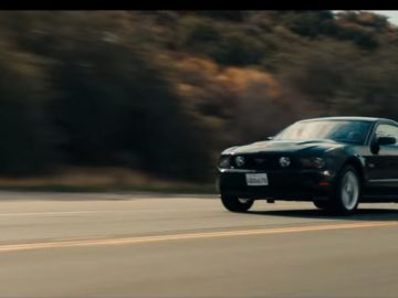 Naast een vrachtwagen rijdt een zwarte Mustang over de weg.