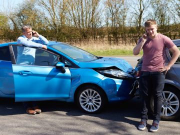 Twee mannen staan naast een blauwe auto met een kapotte voorruit het pamflet van het Verbond van Verzekeraars te raadplegen.