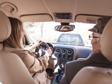 Een vrouw bestuurt een auto met een oudere man, de eigenaar, op de achterbank.