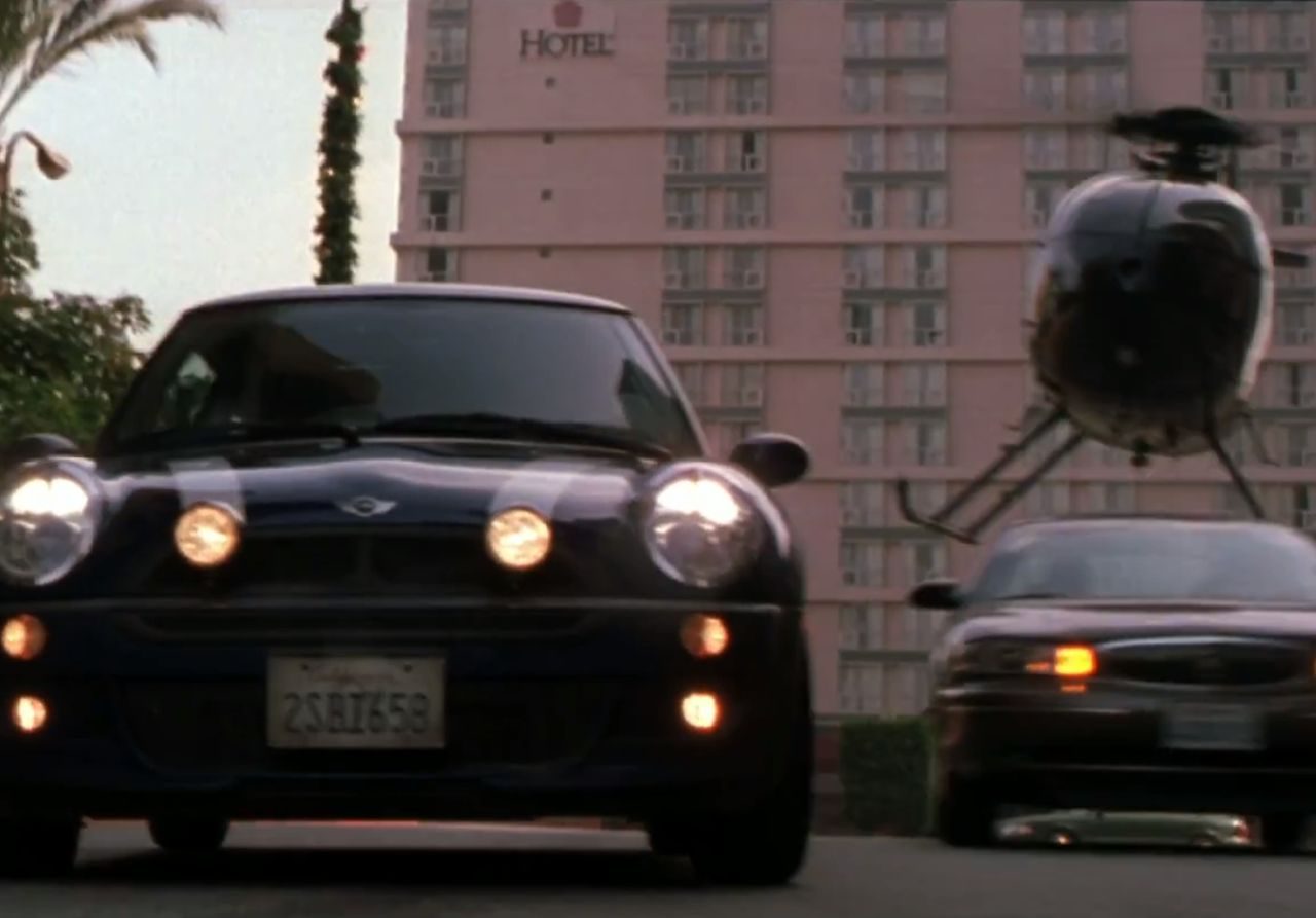 Een blauwe mini cooper van The Italian Job die door de straat rijdt met een helikopter op de achtergrond.