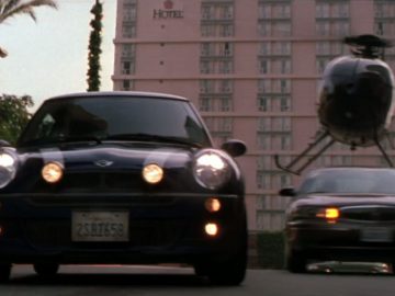 Een blauwe mini cooper van The Italian Job die door de straat rijdt met een helikopter op de achtergrond.