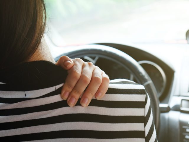 Een vrouw die in een auto zit met haar hand op haar schouder en rugpijn ervaart.