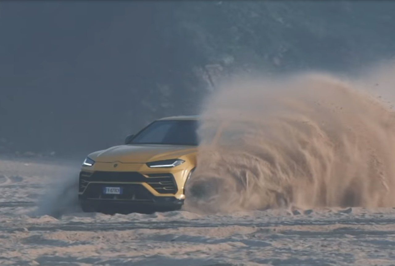 Een gele Urus die door zand op een strand rijdt.