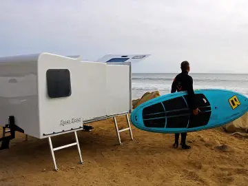 Een man met een surfplank die naast een kleine camper met trekhaak staat.