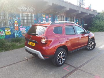 Een rode Dacia Duster TCe 130 geparkeerd voor een met graffiti bedekt gebouw.