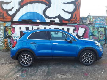Een blauwe Fiat 500X Opening Edition SUV geparkeerd voor een met graffiti bedekte muur.