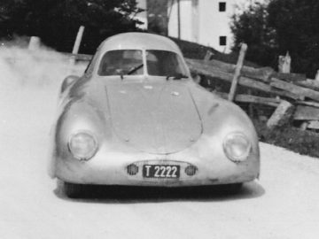 Een oude foto van een Porsche Type 64 die over een onverharde weg rijdt.