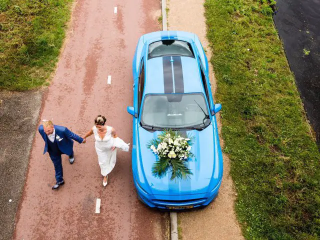 Een bruid en bruidegom lopen naast een blauwe Mustang-sportwagen.