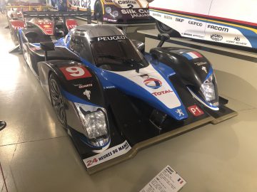 In een museum staat een blauw-witte Le Mans-racewagen tentoongesteld.