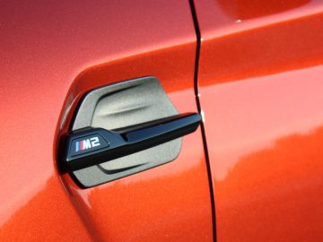 Een close-up van de deurklink van een oranje BMW M2 Competition.
