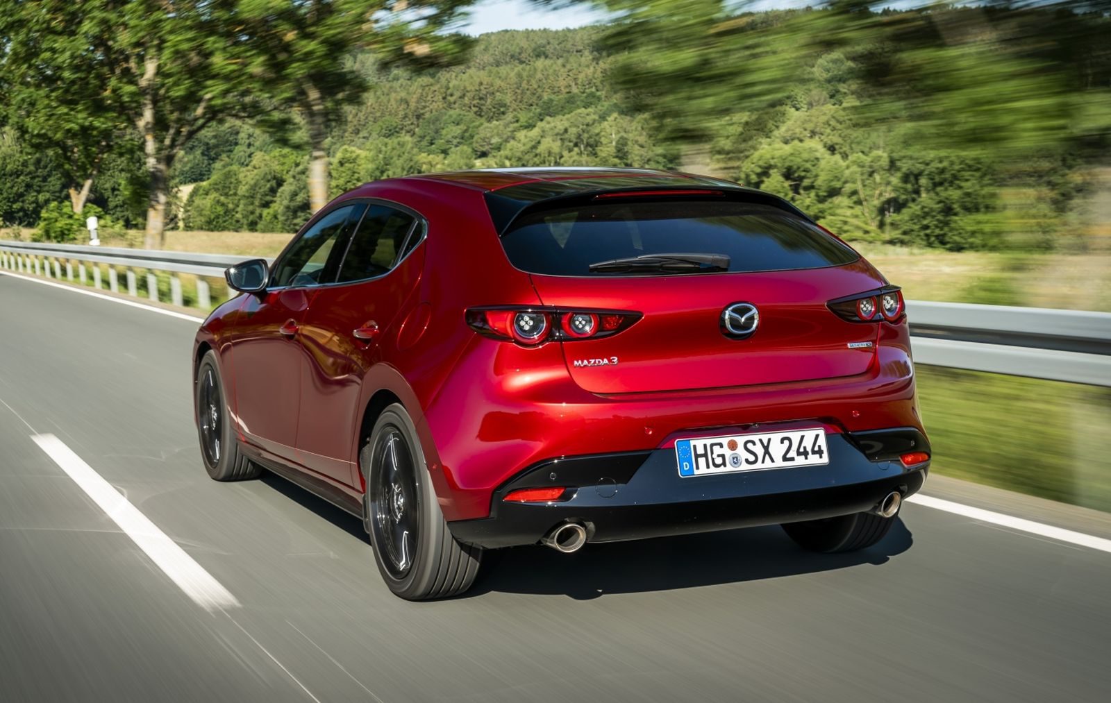 De achterkant van een rode Mazda 3 hatchback die over een weg rijdt.