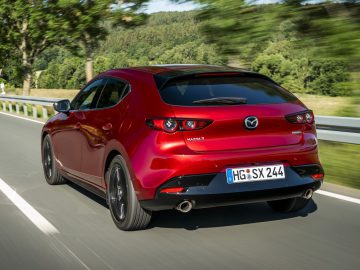 De achterkant van een rode Mazda 3 hatchback die over een weg rijdt.