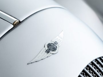 Een close-up van het Morgan Plus 4-embleem op een zilveren sportwagen.