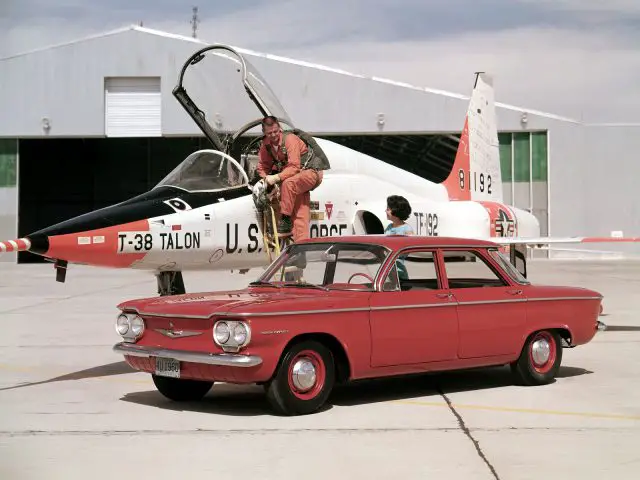 Een man die naast een rode Chevrolet Corvair naast een straaljager staat.