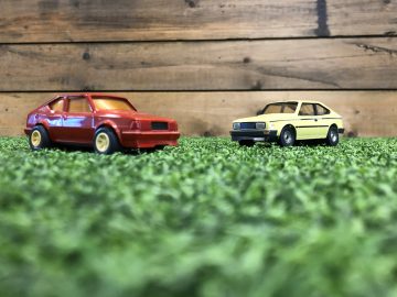Twee Skoda Rapid Coupé speelgoedauto's op gras naast een houten muur.
