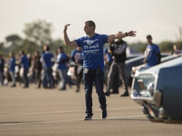 Een man in een blauw T-shirt zwaait met zijn armen voor een menigte met daarop een Ford Mustang.
