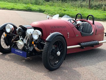 Een rood-zwarte Morgan 3 Wheeler staat geparkeerd aan de kant van de weg.