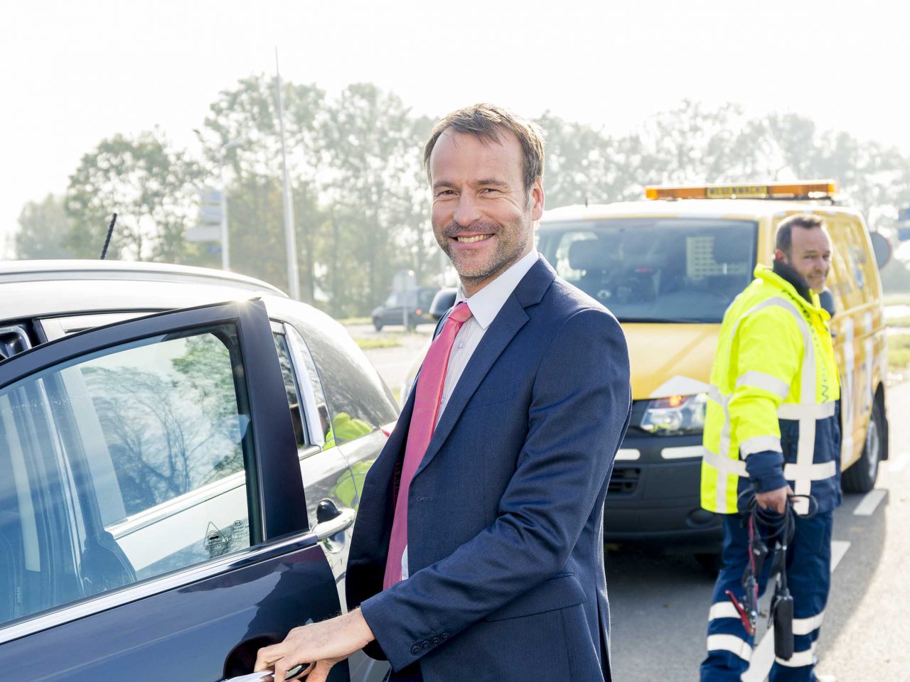 Een man in pak staat naast een auto, wachtend op pechhulp.