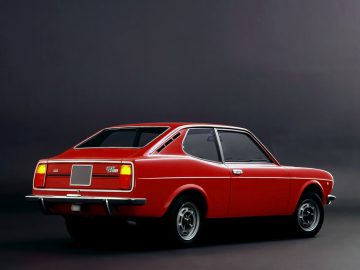 Een rode Fiat 128 staat geparkeerd op een grijze achtergrond.