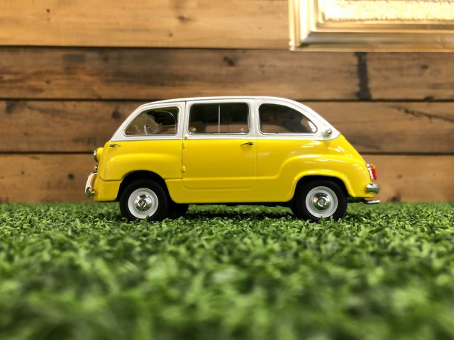 Een gele Fiat 600 Multipla-speelgoedauto staat op het gras voor een houten muur.