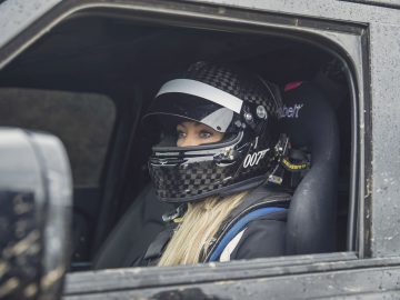 Een vrouw, die lijkt op een personage uit een James Bond-film, met een helm op de bestuurdersstoel van een voertuig.
