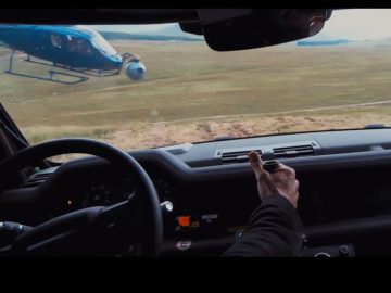 Een man, die op James Bond lijkt, bestuurt een auto met een helikopter op de achtergrond.
