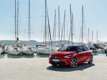 Een rode Opel Corsa geparkeerd voor een jachthaven met boten.