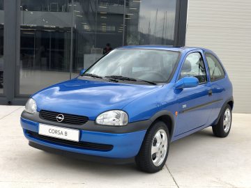 Een kleine blauwe Opel Corsa geparkeerd voor een gebouw.