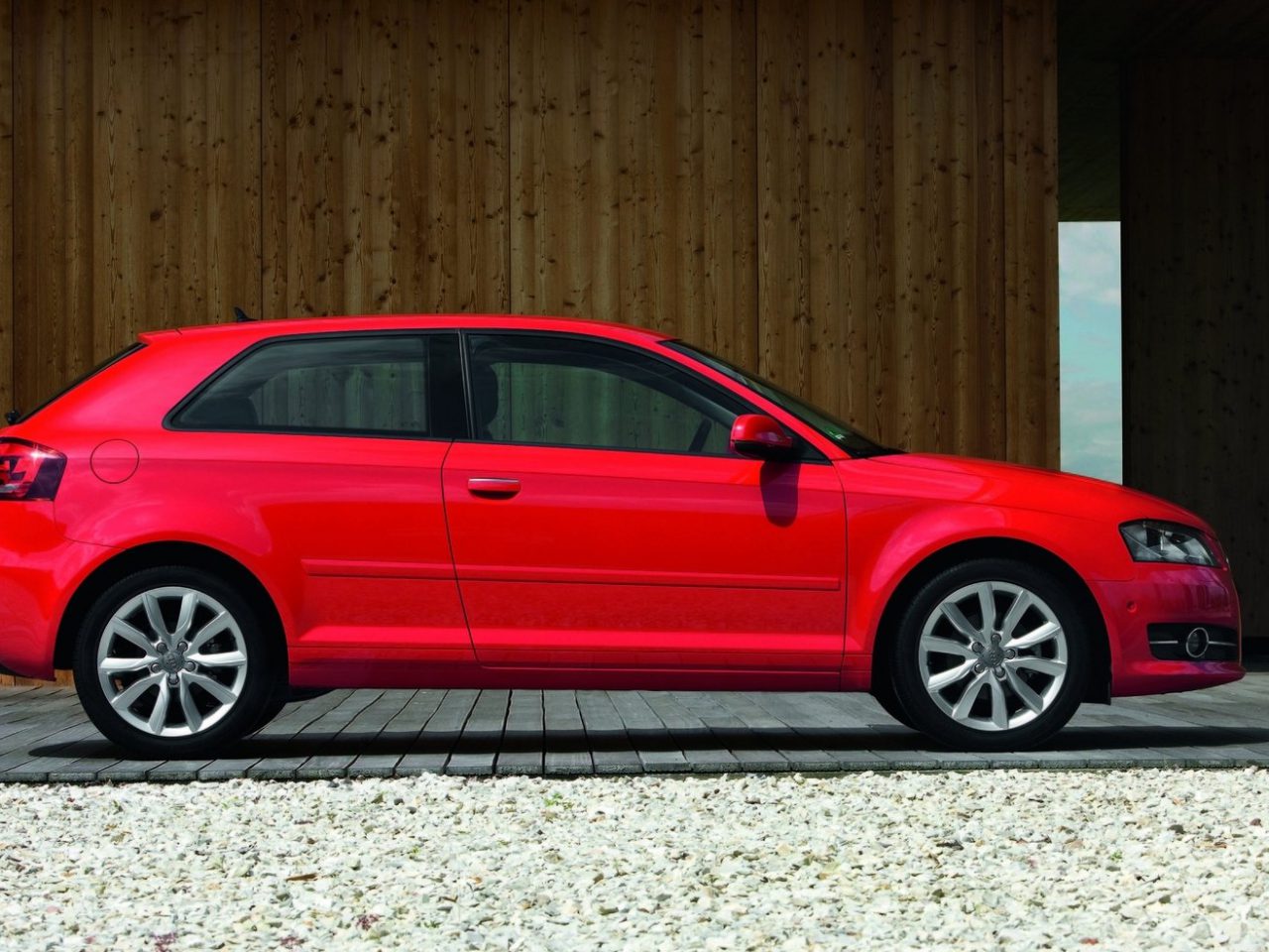 Een rode Audi A3 met een gemiddelde leeftijd is geplaatst voor een houten gebouw.