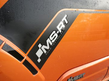 Close-up van een oranje Ford Transit Custom met het "ms-rt"-logo, regendruppels zichtbaar op het oppervlak.