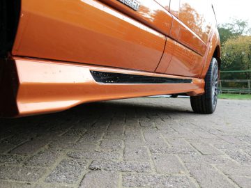Close-up van het oranje zijpaneel en de onderkant van een Ford Transit Custom MS-RT, waarbij de zijskirt wordt benadrukt, geparkeerd op een verhard oppervlak.