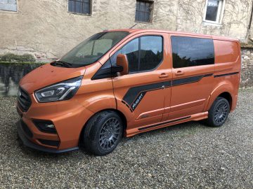 Oranje Ford Transit Custom MS-RT-busje geparkeerd op grind tegen een oud gebouw.