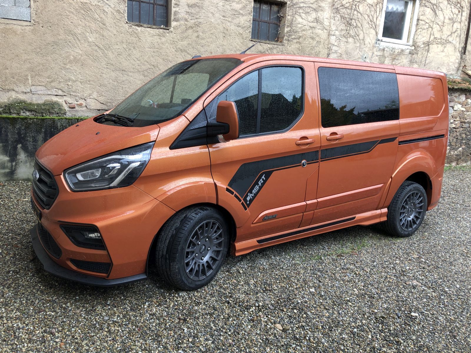 Oranje Ford Transit Custom MS-RT-busje geparkeerd op grind tegen een oud gebouw.